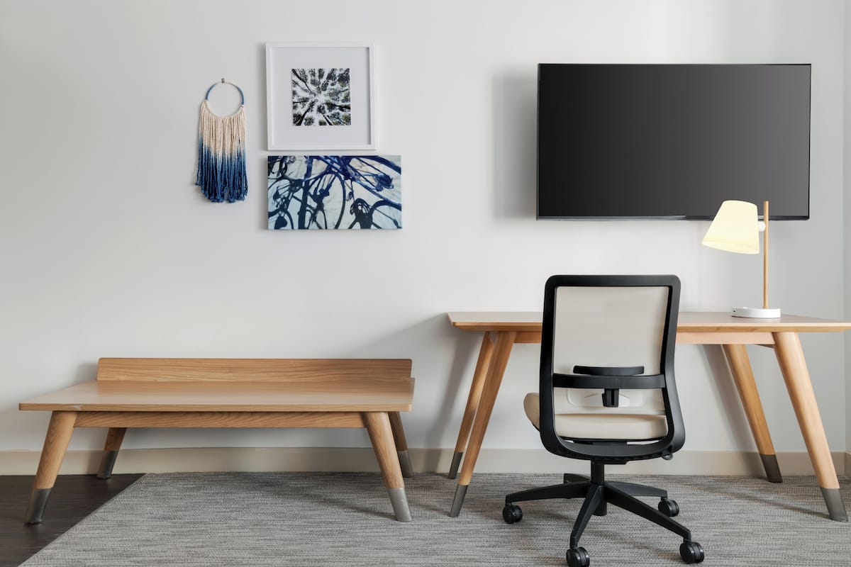 A modern office space with a wooden desk, a black office chair, a bench, a wall-mounted TV, and framed artwork.