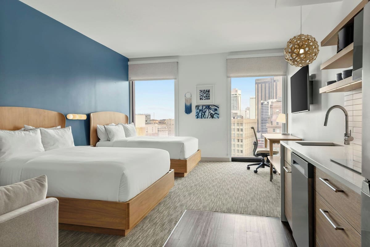 A hotel room with two beds, a desk, a chair, a kitchen area, and a view of the city.