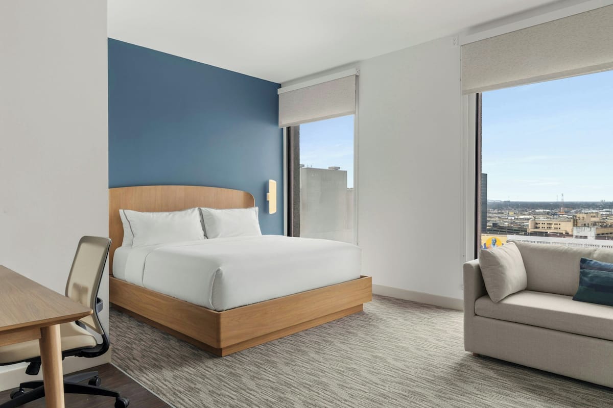A hotel room with a bed, a desk, a sofa, and a large window offering a city view.