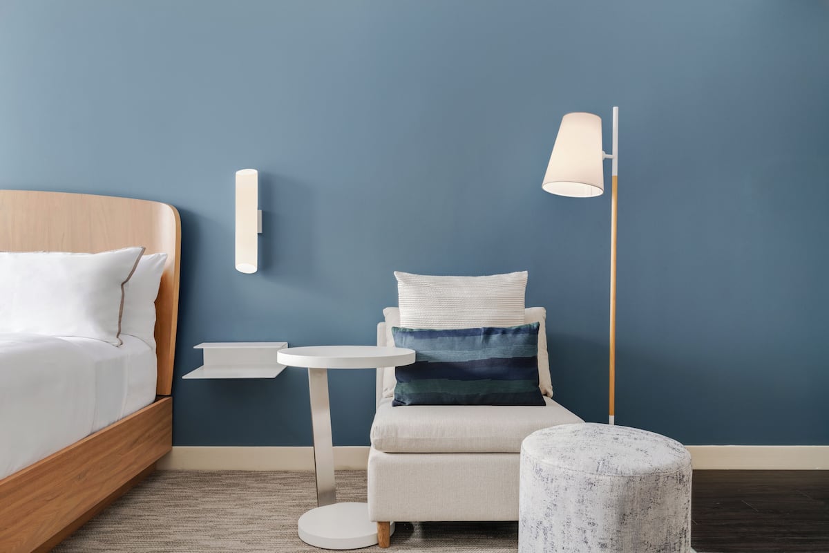 A bedroom with a bed, a bedside table, a chair, a floor lamp, and a small round ottoman.