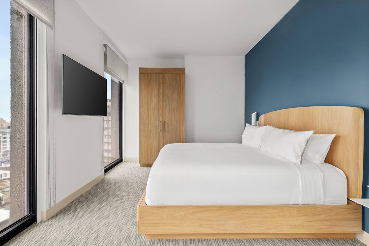 A hotel room with a bed, a TV, a wardrobe, and a view of the city.