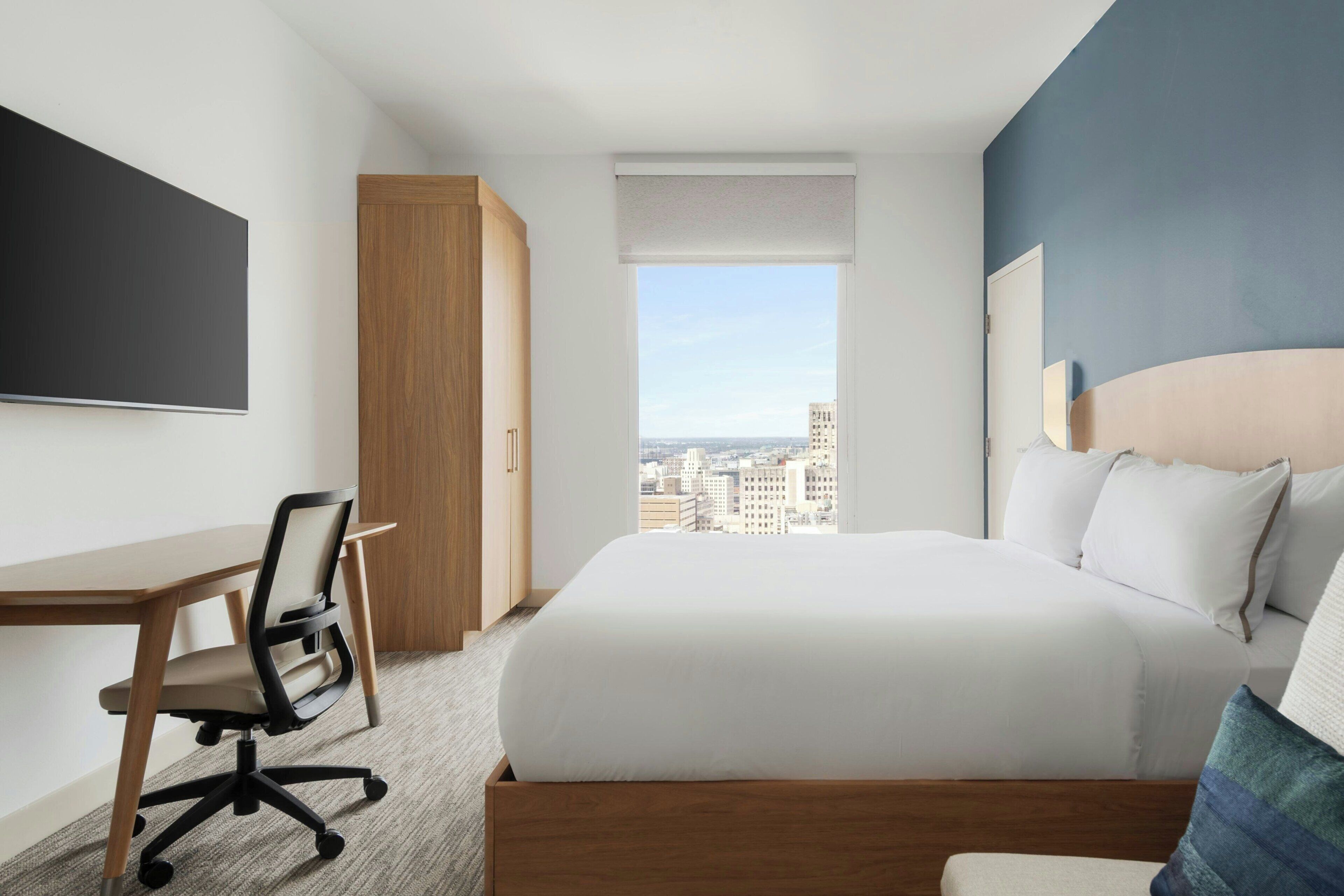 Ein Hotelzimmer mit Bett, Schreibtisch, Stuhl, großem Fenster mit Stadtblick und einem an der Wand befestigten Flachbildfernseher.