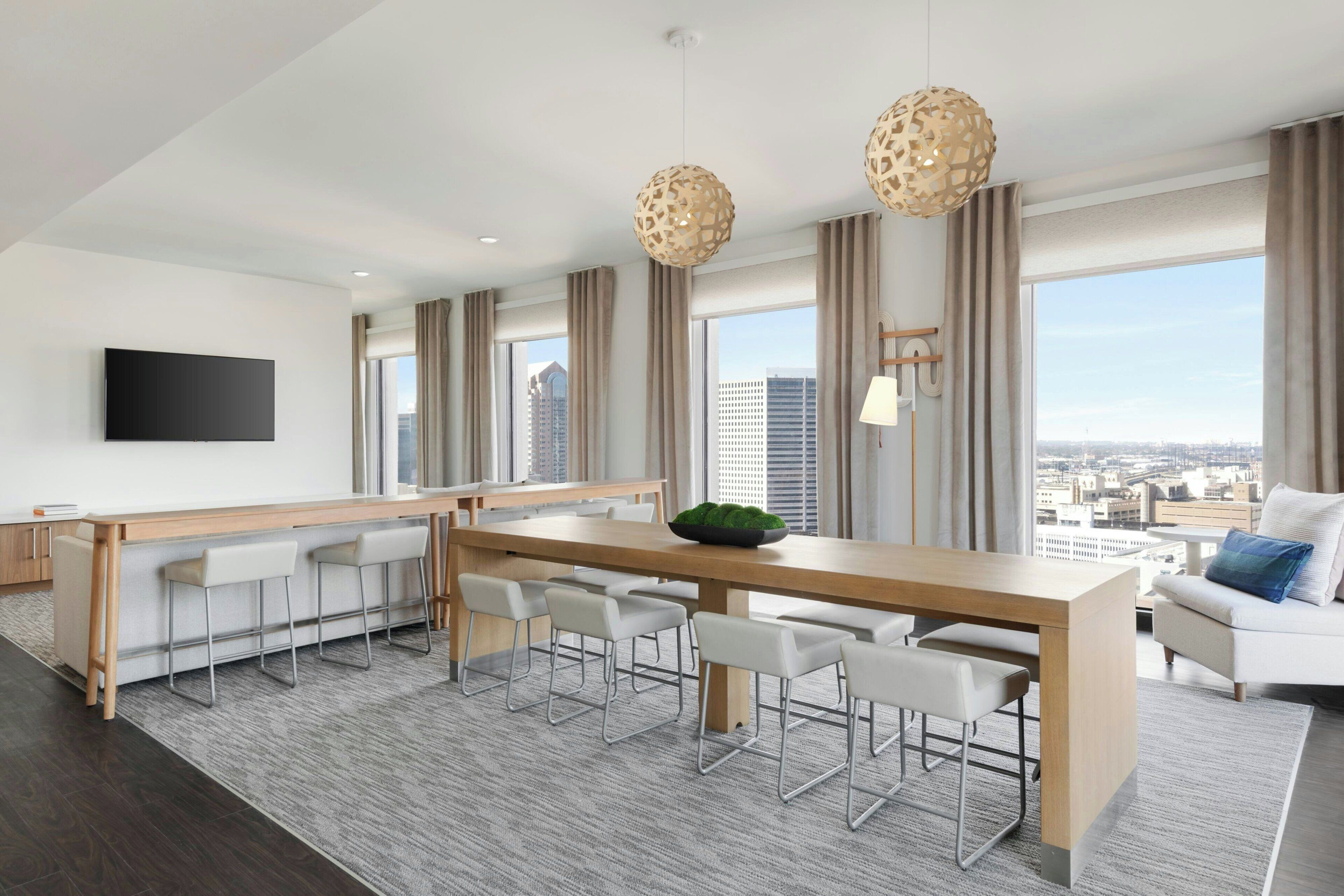 A modern hotel room with a large window, a wooden dining table, white chairs, a flat-screen TV, and a sofa with a view of the city.