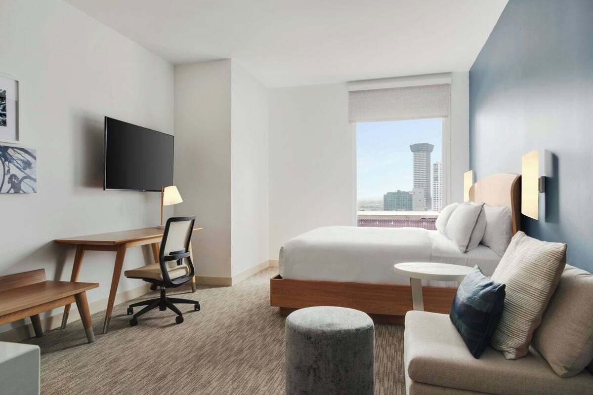 A hotel room with a bed, a desk, a chair, a TV, a window with a city view, and a small round ottoman.