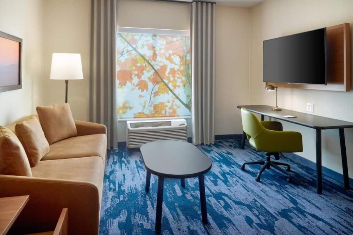 Habitación de hotel con sofá, una mesita, escritorio con silla, televisor de pantalla plana y ventana grande con cortinas.