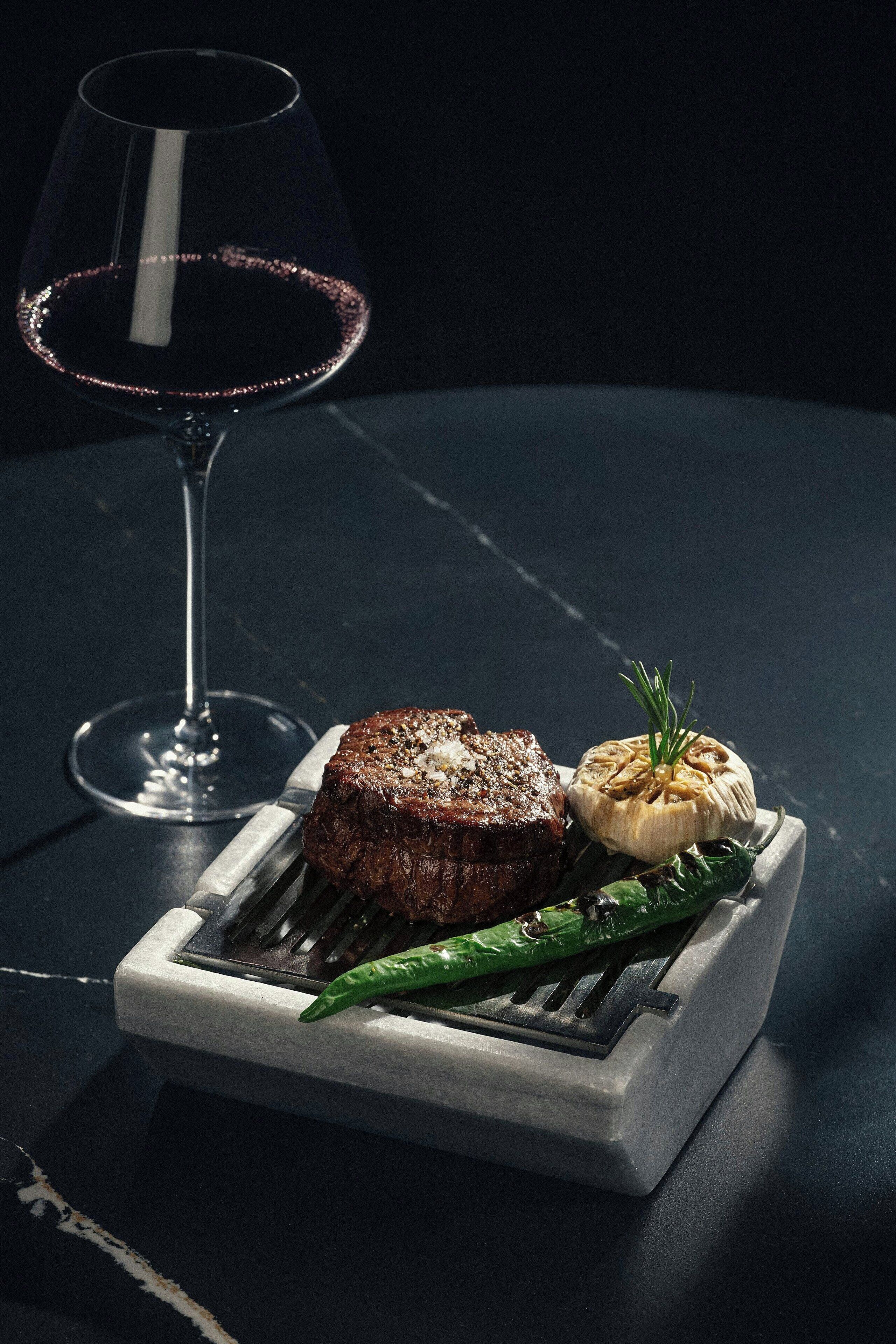 A steak with a side of garlic and a green chili, a glass of red wine, and a metal fork on a dark surface.