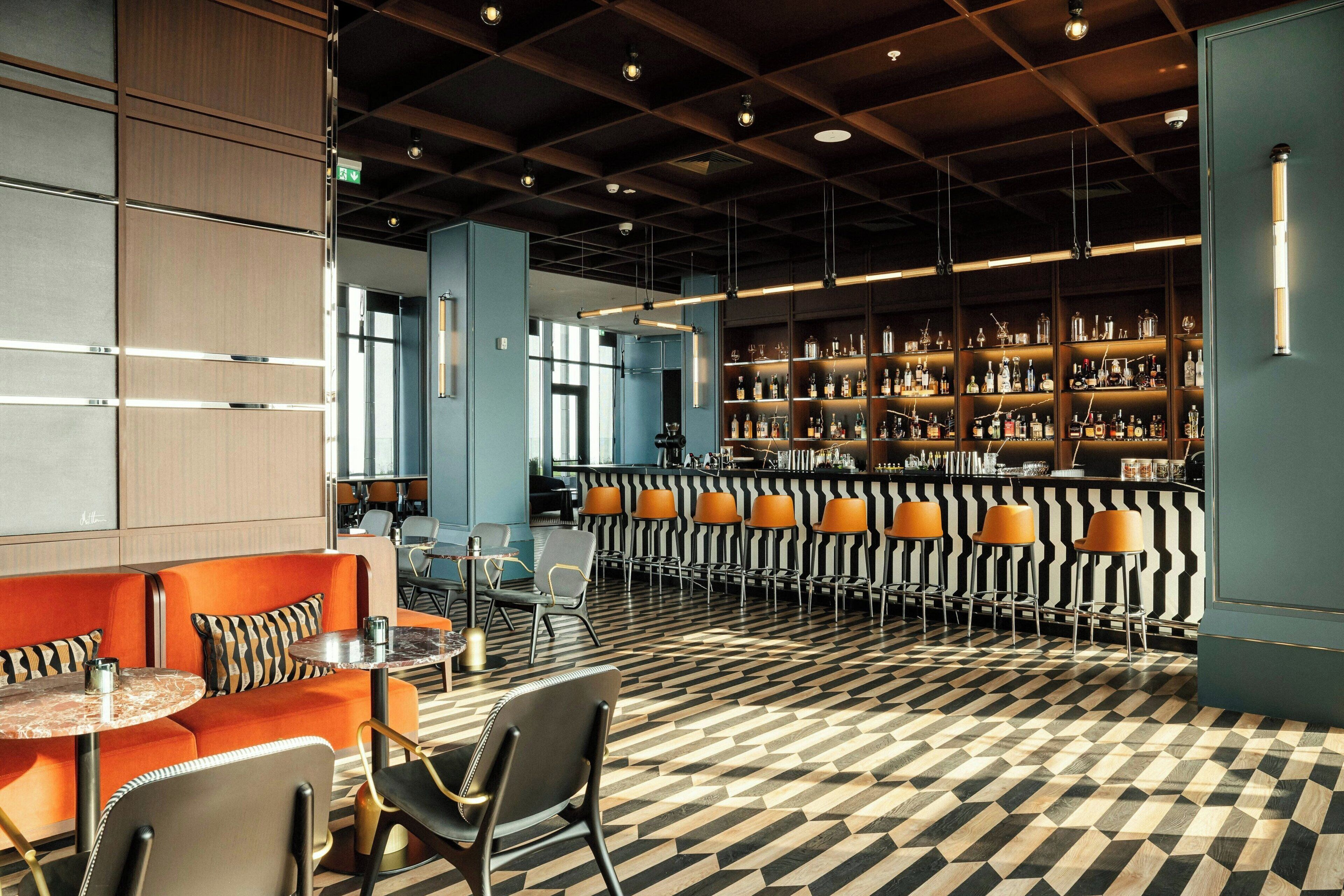 A bar area with a patterned floor, seating with orange and black cushions, and shelves stocked with bottles.