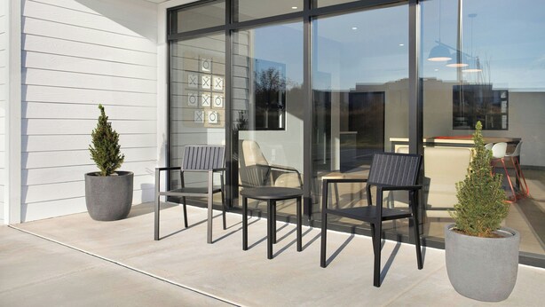 An outdoor seating area with modern chairs and potted plants, a glass wall, and a view of the interior.