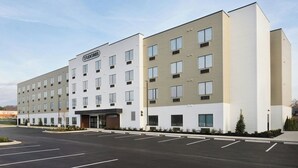 A multi-story hotel with a modern design, featuring a central entrance, multiple windows, and a parking lot in front.