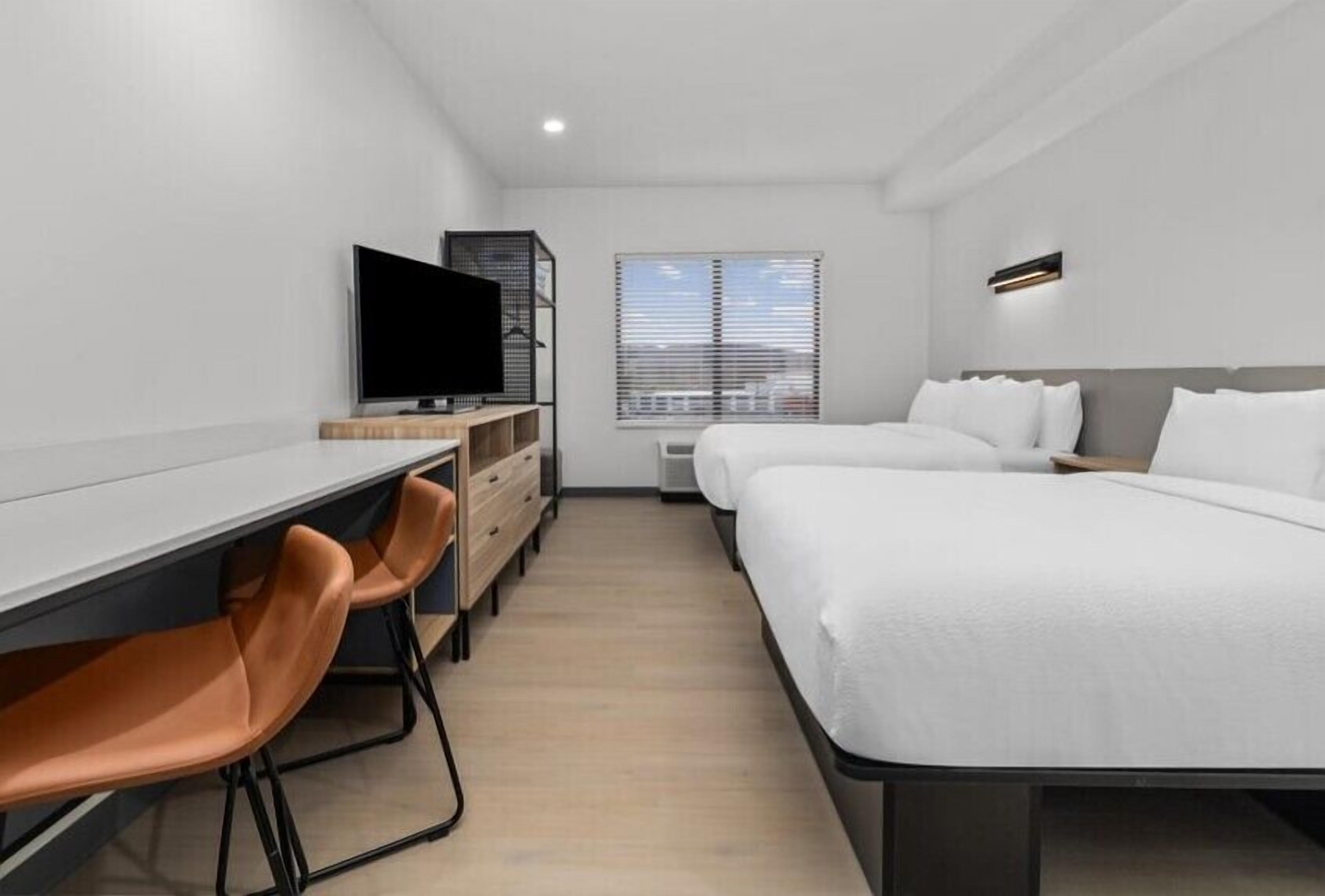 Une chambre d’hôtel moderne équipée d’un bureau, de deux lits, d’une télévision et d’une fenêtre avec des stores.