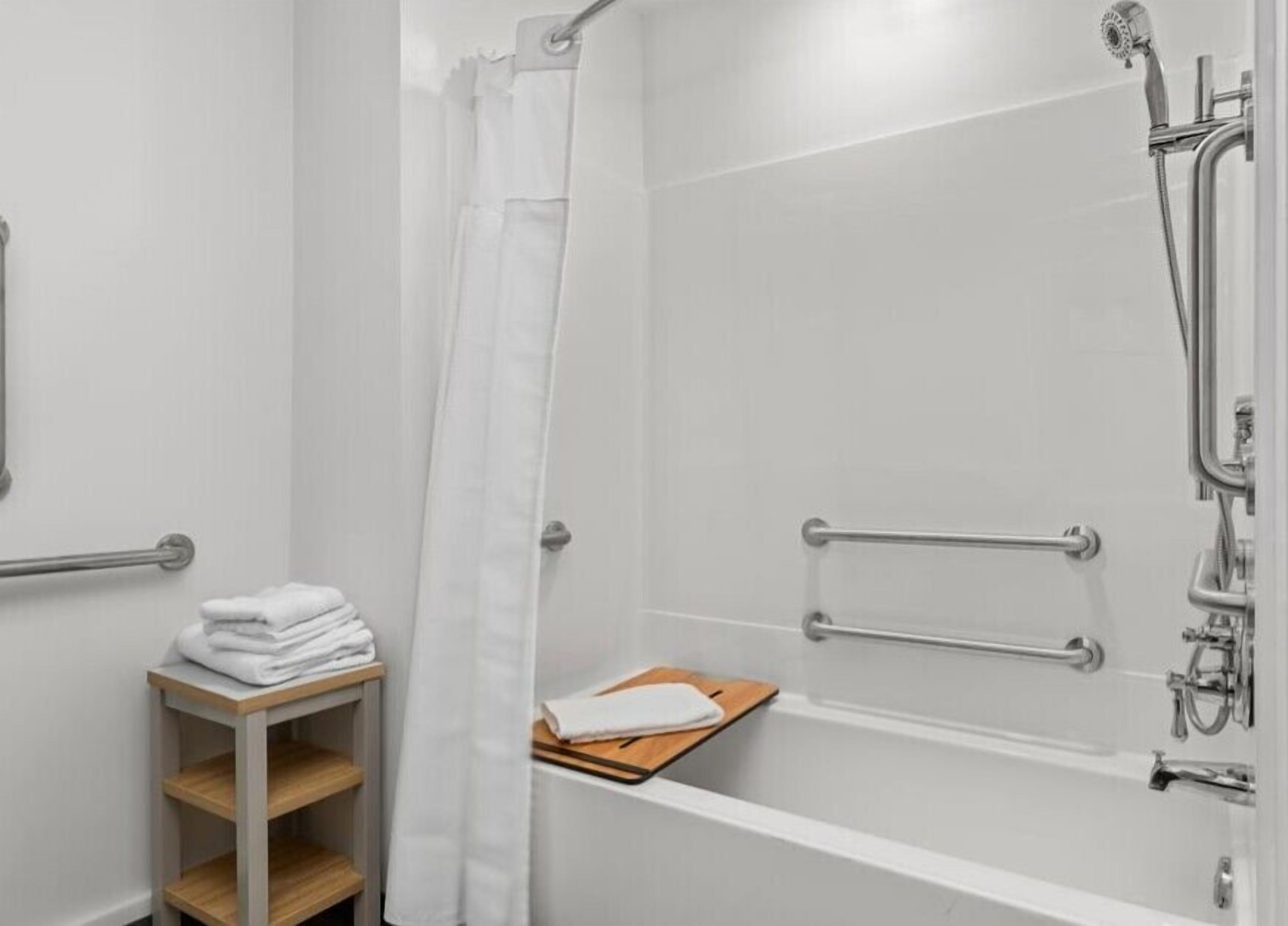 A modern bathroom with a walk-in shower, grab bars, a towel rack, and a small wooden shelf.
