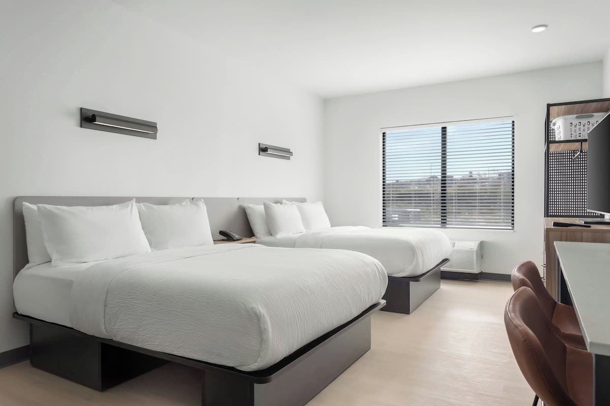 A modern hotel room with two beds, a desk, and a view of the city through large windows.