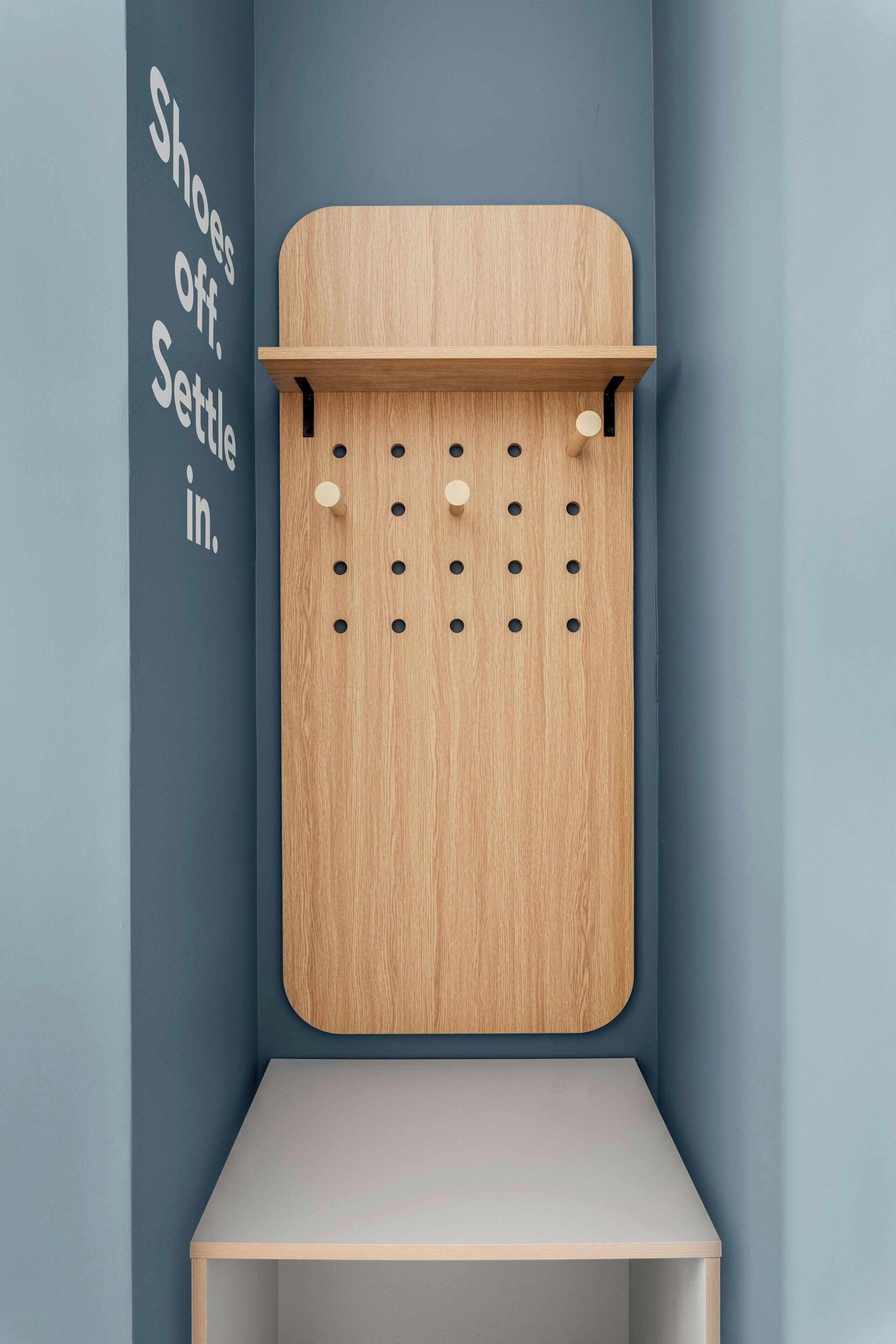 Un coin avec un mur sur lequel est fixé un porte-chaussures en bois et une étagère, et un autre mur sur lequel on peut lire : « Enlevez vos chaussures. Installez-vous confortablement. »