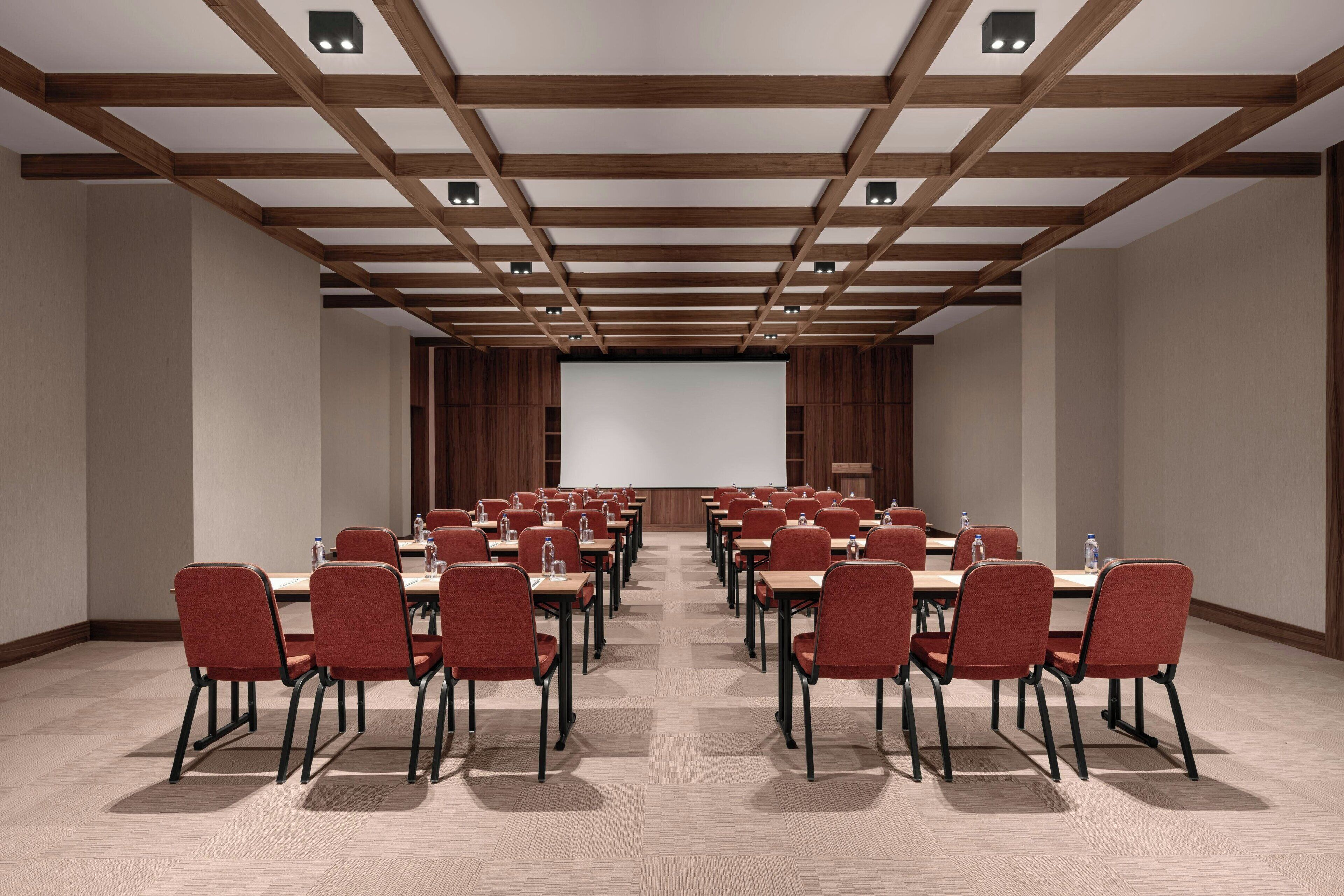 A conference room with rows of red chairs and tables, wooden ceiling beams, and a large screen at the front.