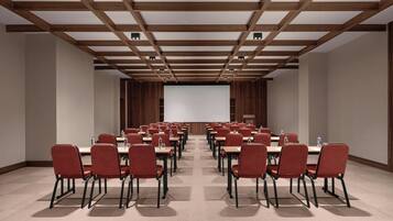 A conference room with rows of red chairs and tables, wooden ceiling beams, and a large screen at the front.