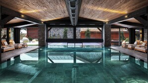 Une vaste piscine intérieure offrant une vue dégagée sur l’extérieur, des poutres en bois au plafond et des espaces de détente de part et d’autre.