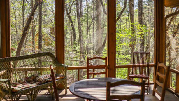 A wooden deck with a table and chairs, surrounded by large windows offering a view of a forest. - Splendor Mountain Ruby Cabin (Tiger)