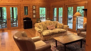 A cozy cabin interior with a wooden floor, a floral sofa, leather armchairs, and large windows offering a view of the outdoors. - Splendor Mountain Ruby Cabin (Tiger)