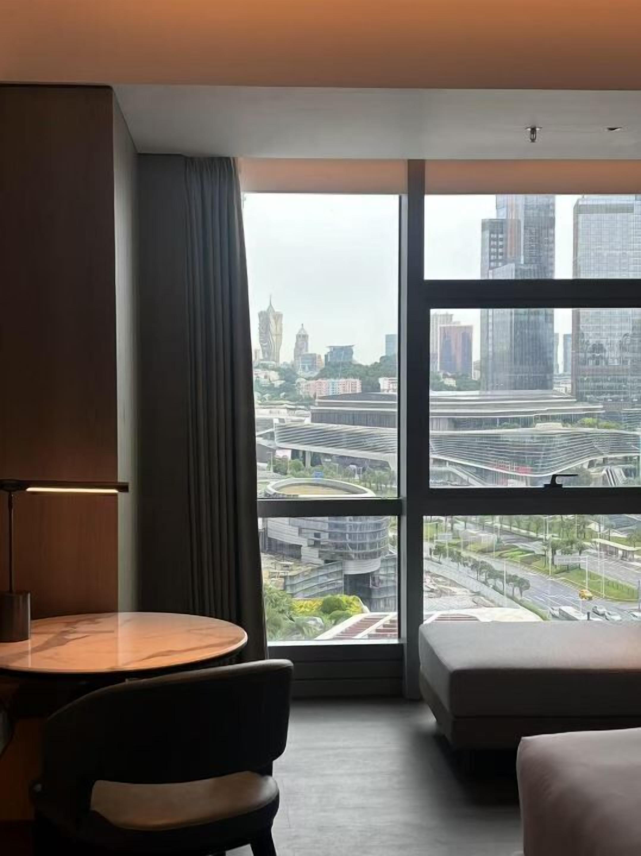 A modern hotel room with a large window offering a city view, a round table, a chair, and a sofa.