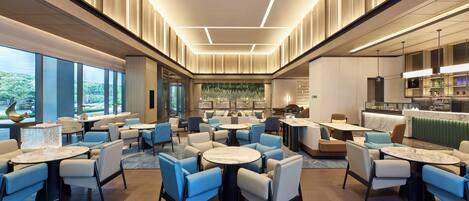 A modern hotel lobby with round tables and blue chairs, large windows, and a bar area.
