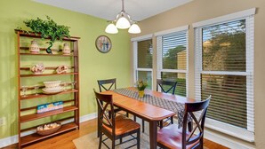 Un coin repas avec une table et des chaises en bois, un lustre, un mur vert et une fenêtre avec des stores.
