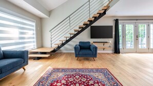 Ein modernes Wohnzimmer mit Holzboden, einem blauen Sofa, einem blauen Sessel, einem großen Teppich in Rot und Blau sowie einer Treppe mit Glastrennwand und einem an der Wand befestigten Flachbildfernseher.