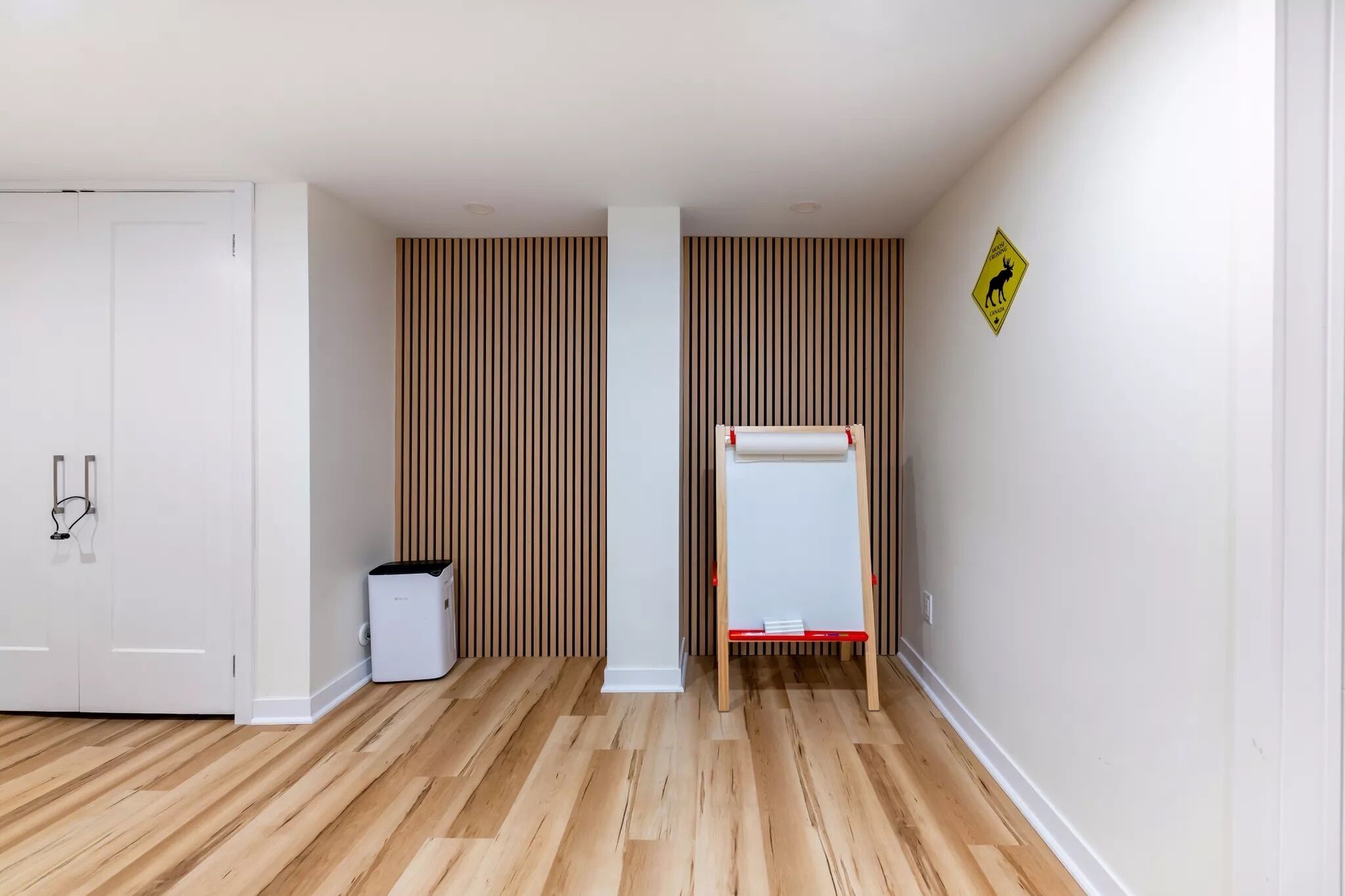A room with wooden flooring, a white door, a whiteboard on a stand, and a trash bin.