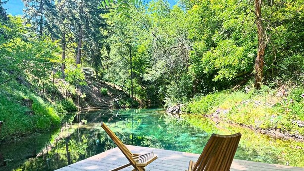 Une terrasse en bois surplombant une rivière aux eaux claires, avec une végétation luxuriante et des arbres en arrière-plan.