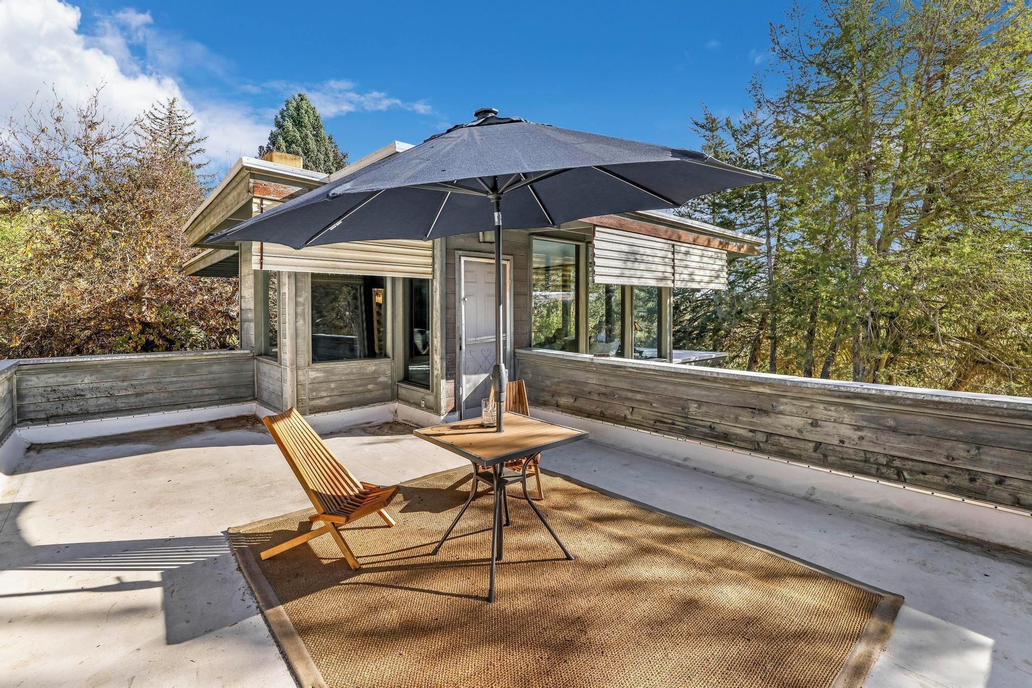 A modern outdoor patio with a table and chairs, a large umbrella, and a view of a contemporary house.