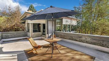 A modern outdoor patio with a table and chairs, a large umbrella, and a view of a contemporary house.