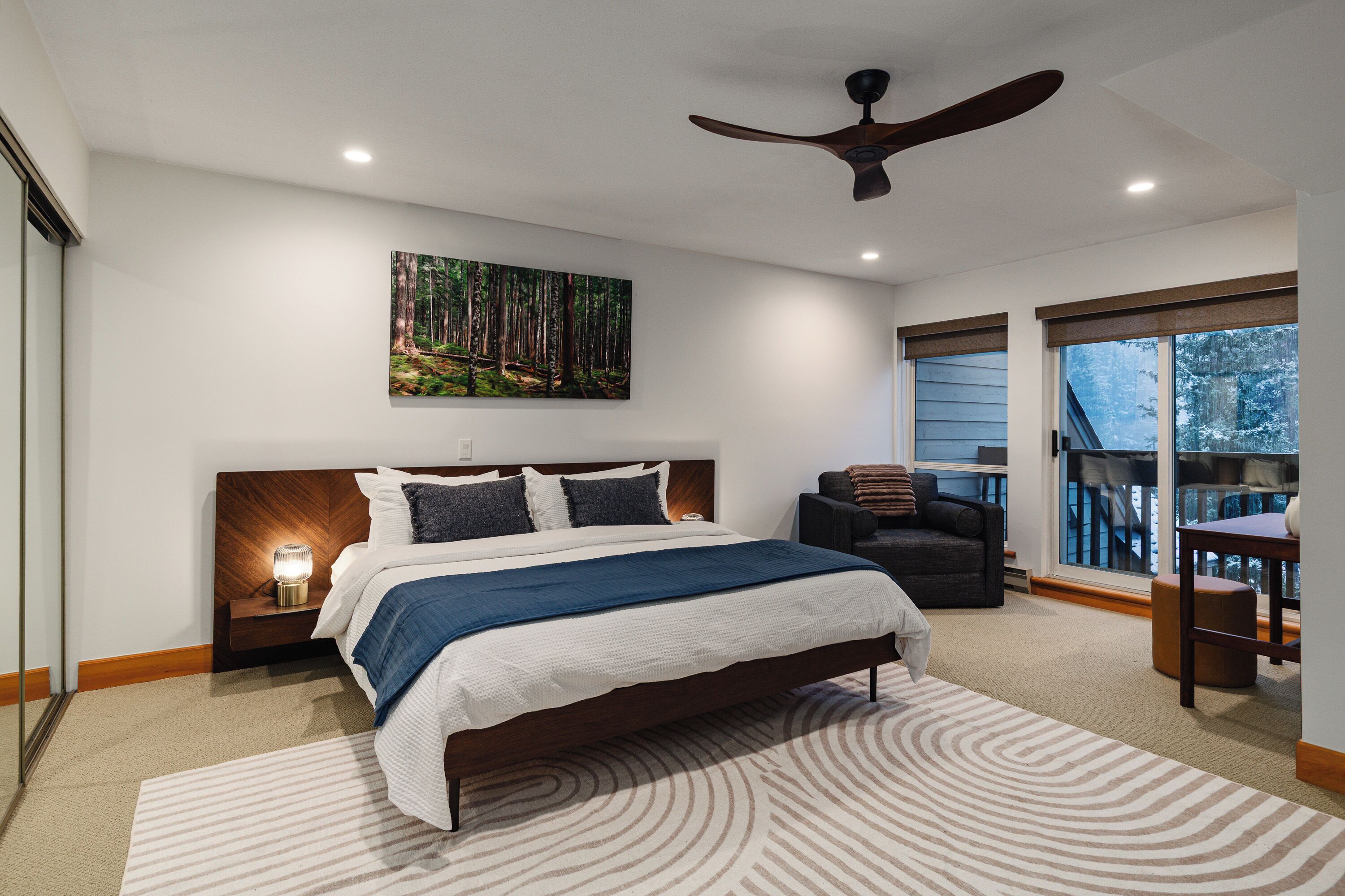 A modern bedroom with a large bed, a ceiling fan, a forest-themed painting, and a view of the outdoors through a window.