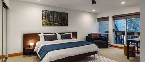 A modern bedroom with a large bed, a ceiling fan, a forest-themed painting, and a view of the outdoors through a window.