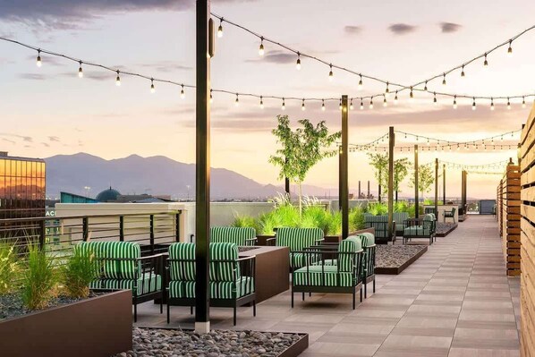 An outdoor seating area with green-striped chairs and tables, string lights overhead, and a view of mountains in the background.