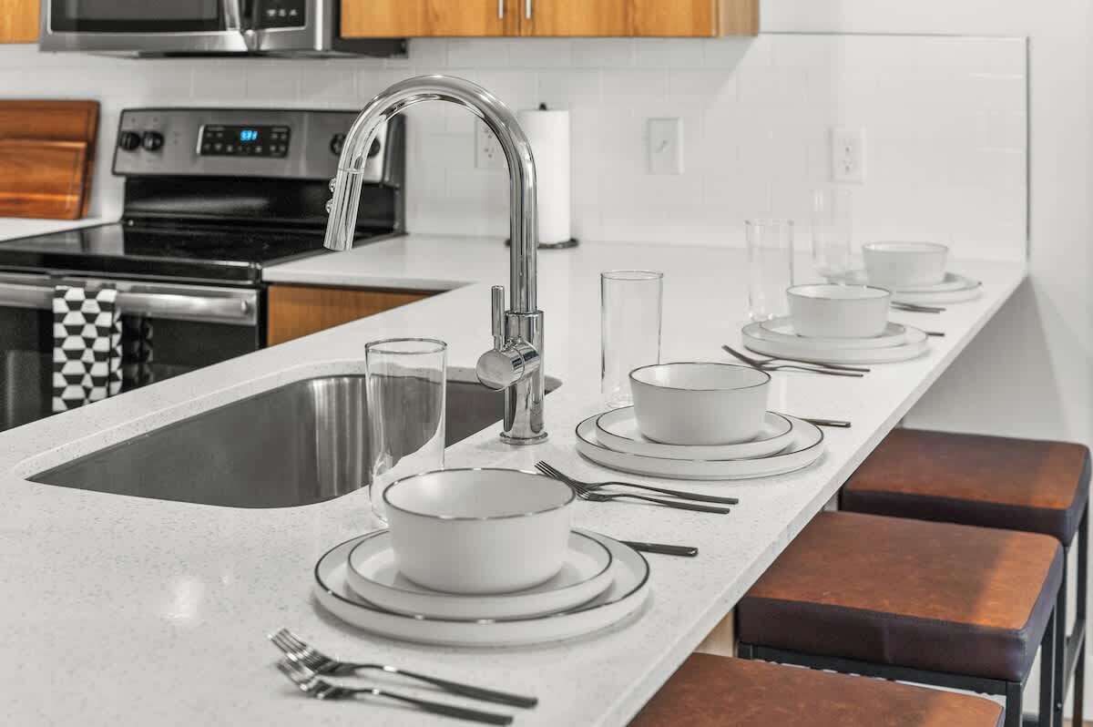 Una cocina moderna con un lavamanos de acero inoxidable, un microondas y una estufa. Hay cuencos y platos blancos sobre la encimera, junto con cubiertos.