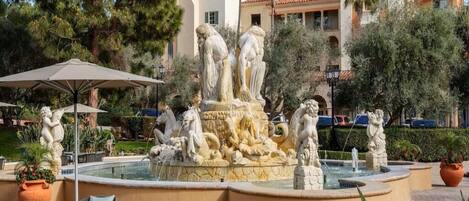 Una fuente con estatuas, rodeada de mobiliario exterior y palmeras.