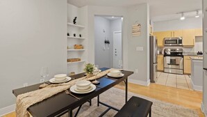 Uma cozinha moderna com eletrodomésticos de aço inoxidável, uma área de jantar com mesa escura e pratos brancos, e um corredor que dá para outro quarto.