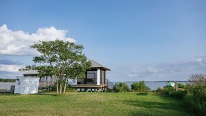 Een vakantiehuis aan het meer met een terras en een kleine schuur, omringd door groen en een heldere lucht.