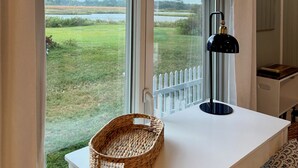 Een kamer met een groot raam dat uitkijkt op een grasveld en een wateroppervlak. Er staat een wit bureau met een gevlochten mand en een zwarte bureaulamp.