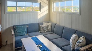 A cozy room with a grey sofa, a wooden table, and a view of a green landscape through two windows.