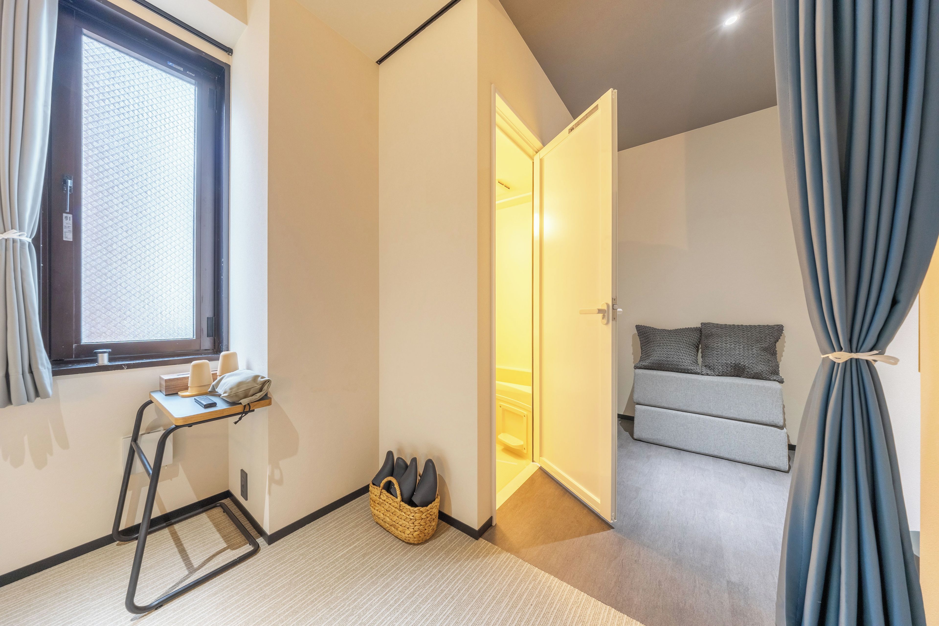 Habitación pequeña con cama, escritorio y silla, ventana con cortinas y puerta que da a otra habitación.