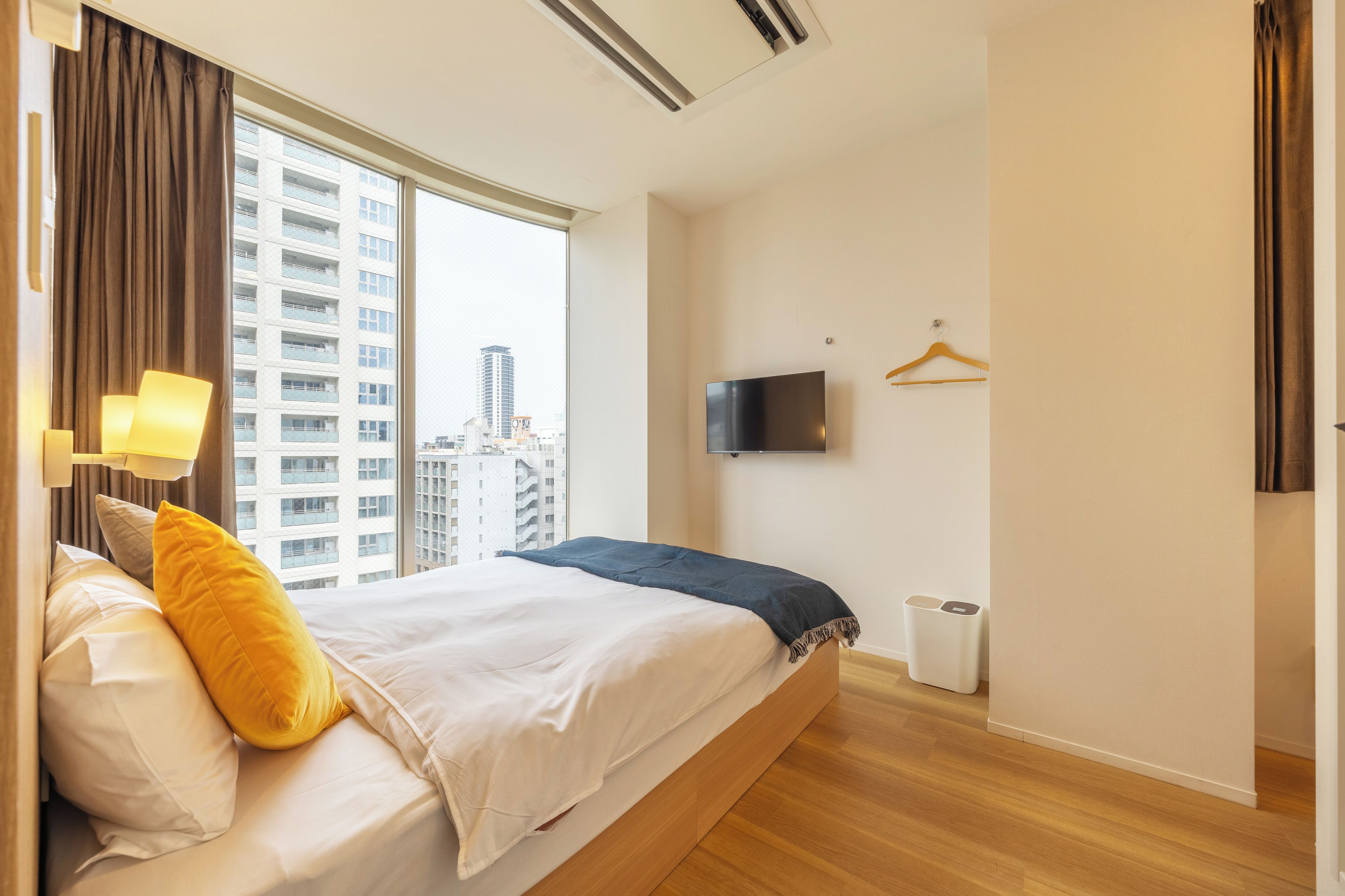 A hotel room with a bed, a TV mounted on the wall, a window with curtains, and a view of buildings outside.