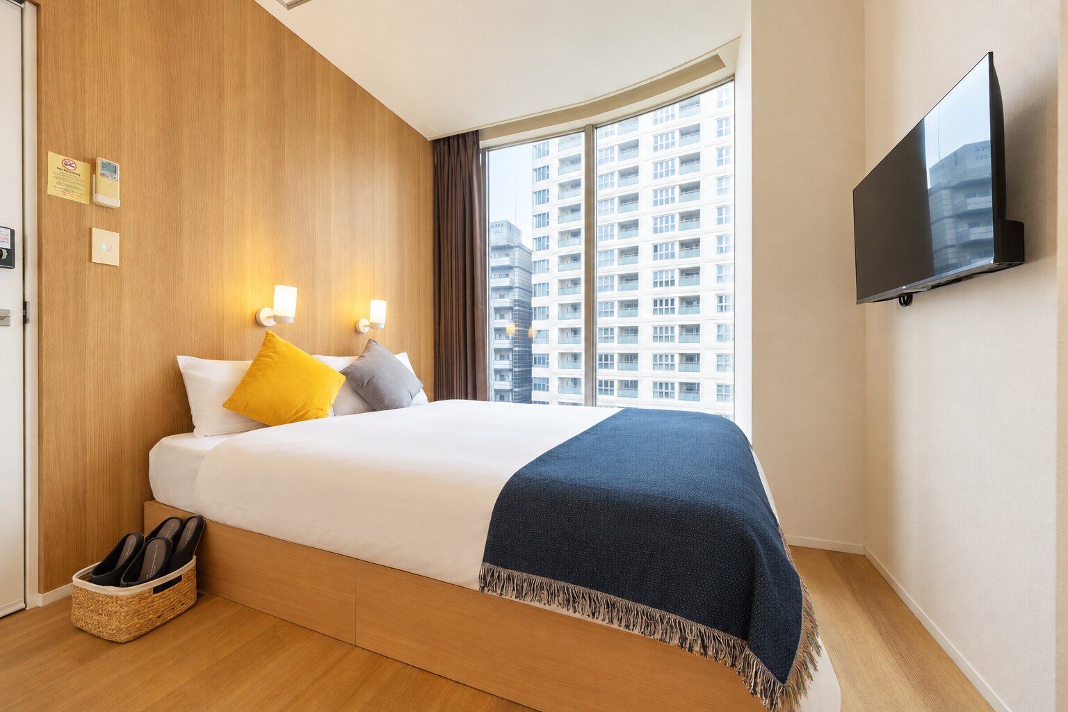 A hotel room with a bed, a TV mounted on the wall, a window with a view of a building, and a shoe rack.