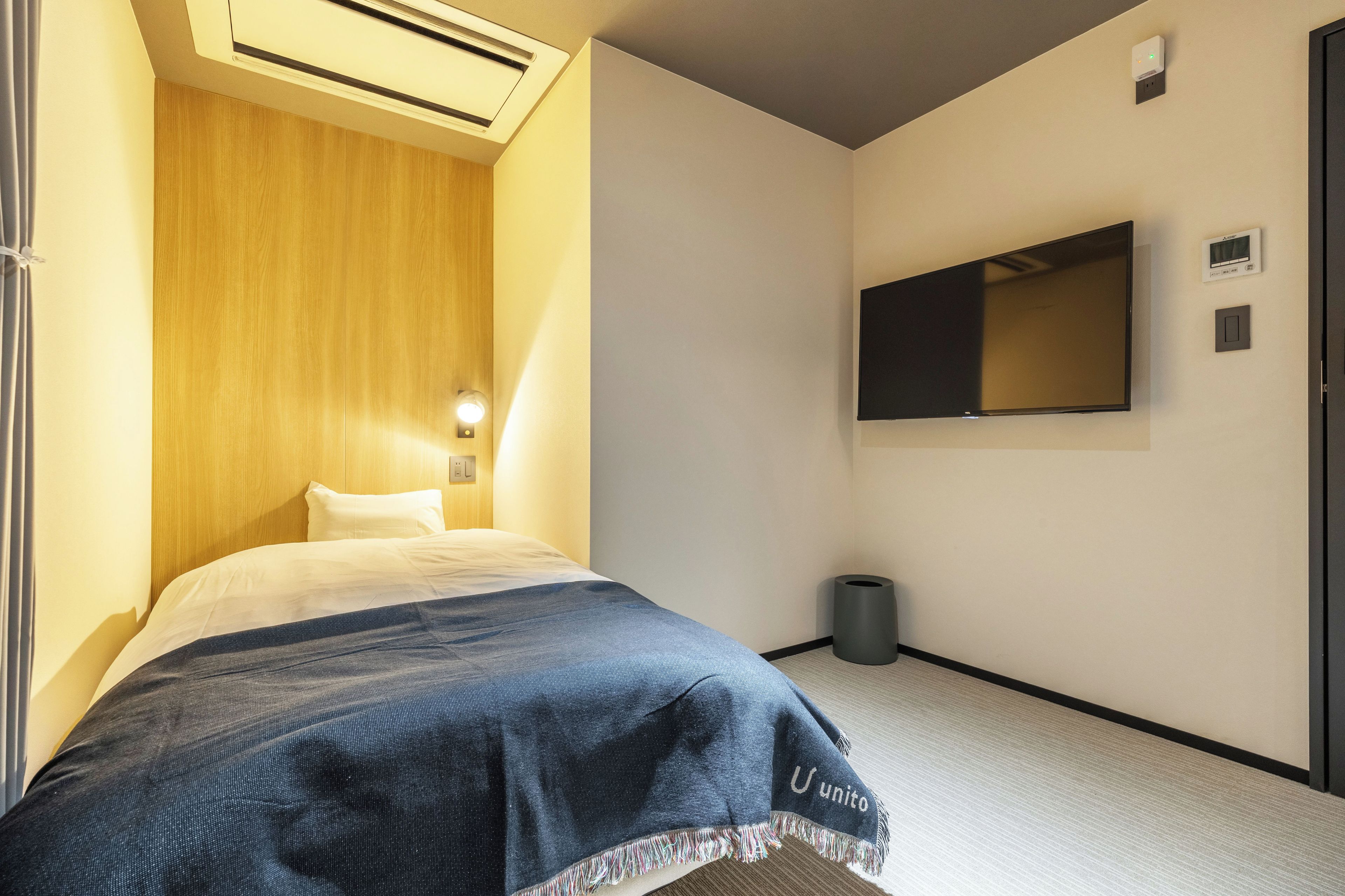 A hotel room with a bed, a wall-mounted TV, a small trash bin, and a ceiling-mounted air vent.