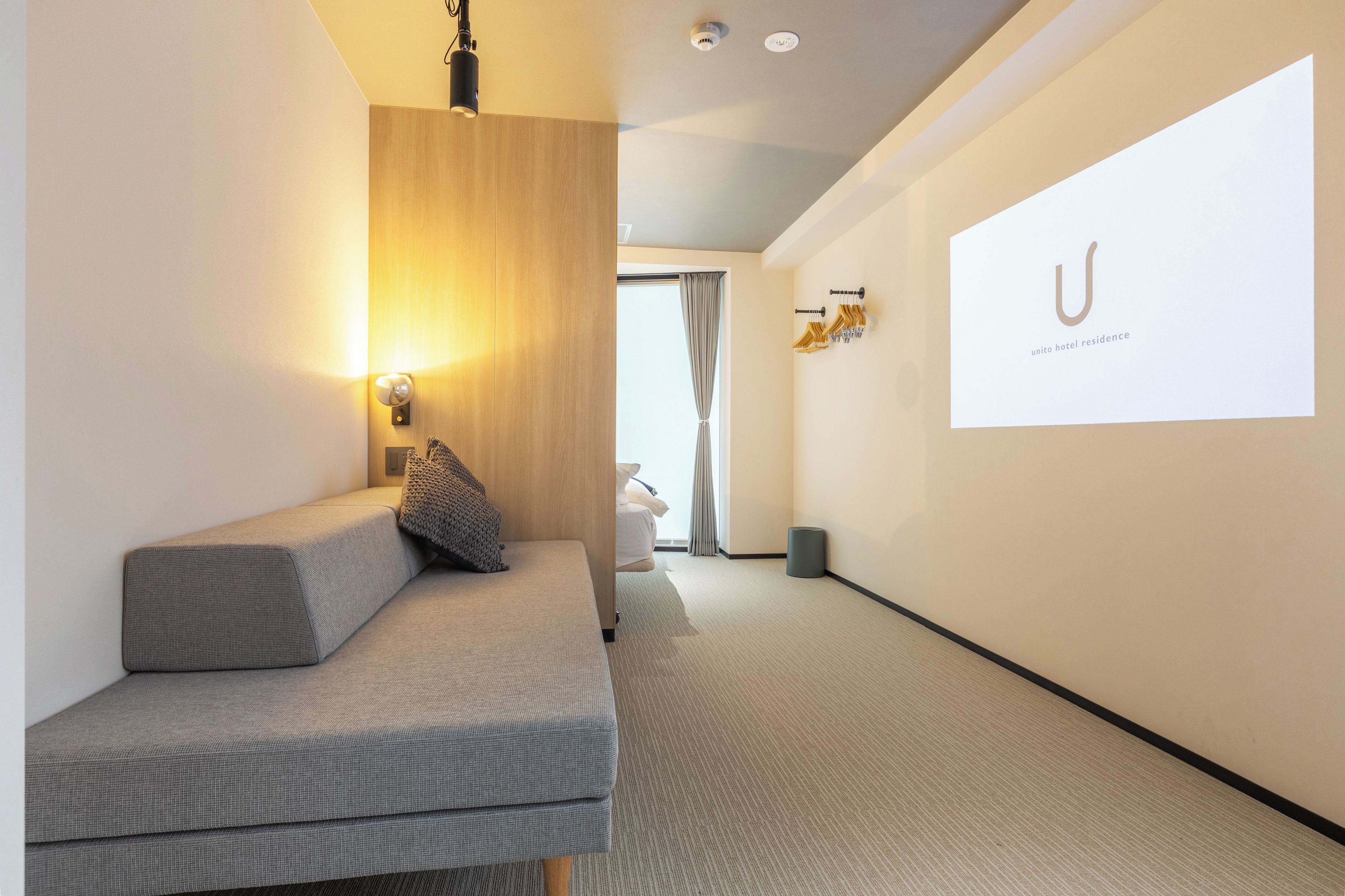 Una habitación de hotel moderna con sofá, una lámpara de pared y una pantalla de proyección mostrando el logotipo del hotel.