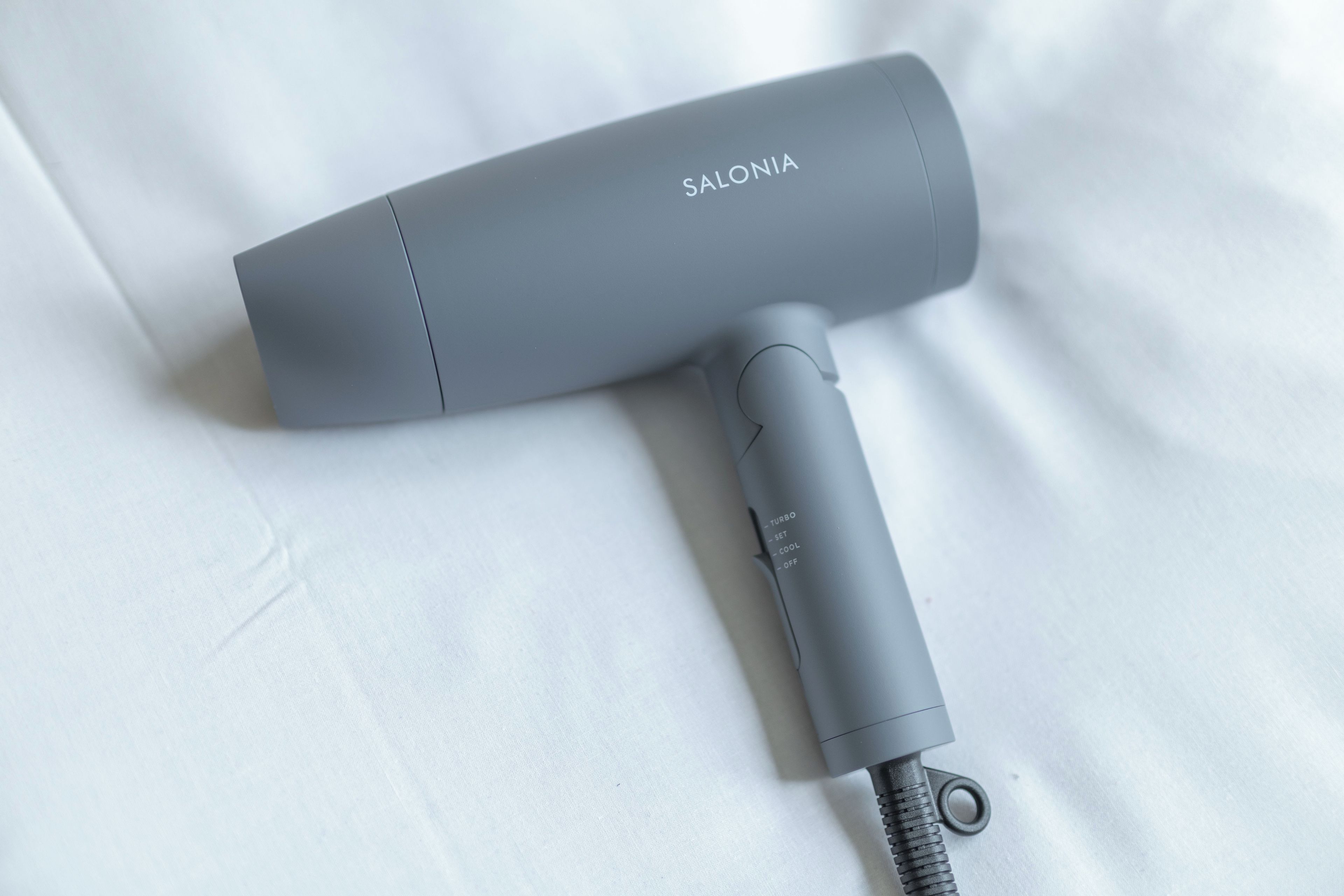 Un sèche-cheveux gris, portant la marque « SALONIA », est posé sur une surface blanche.