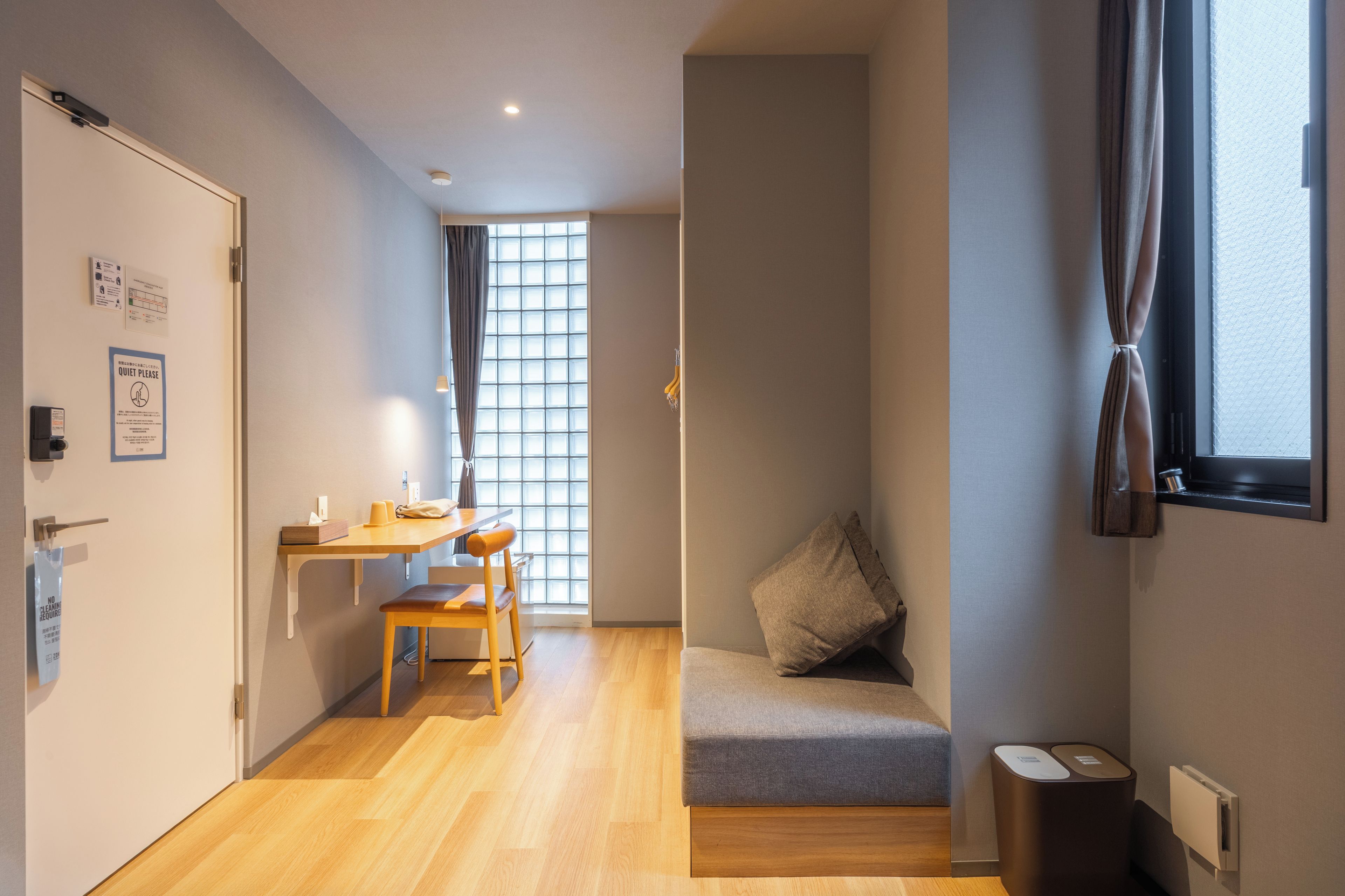 A compact room with a wooden floor, a small table with a chair, a bench with cushions, a window with curtains, and a wall-mounted trash bin.