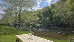 A picnic table with a bottle and glasses, surrounded by lush greenery and a river. - Dog-Kids and Family Friendly Riverfront Cabin! (Crumpler)