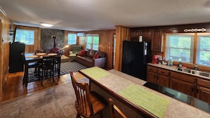 A spacious kitchen with wooden cabinets, a dining area with a table and chairs, a stone accent wall, and a large window with blinds. - Dog-Kids and Family Friendly Riverfront Cabin! (Crumpler)