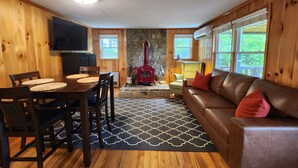 A cozy cabin interior with a wooden dining table, chairs, a leather sofa, a stone fireplace, and large windows. - Dog-Kids and Family Friendly Riverfront Cabin! (Crumpler)