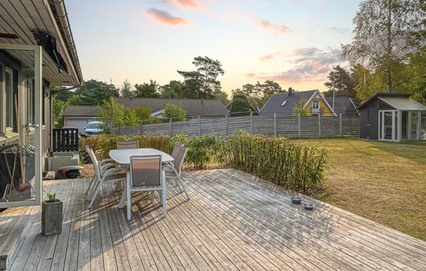 A wooden deck with a table and chairs, surrounded by a fenced yard with trees and a house in the background.